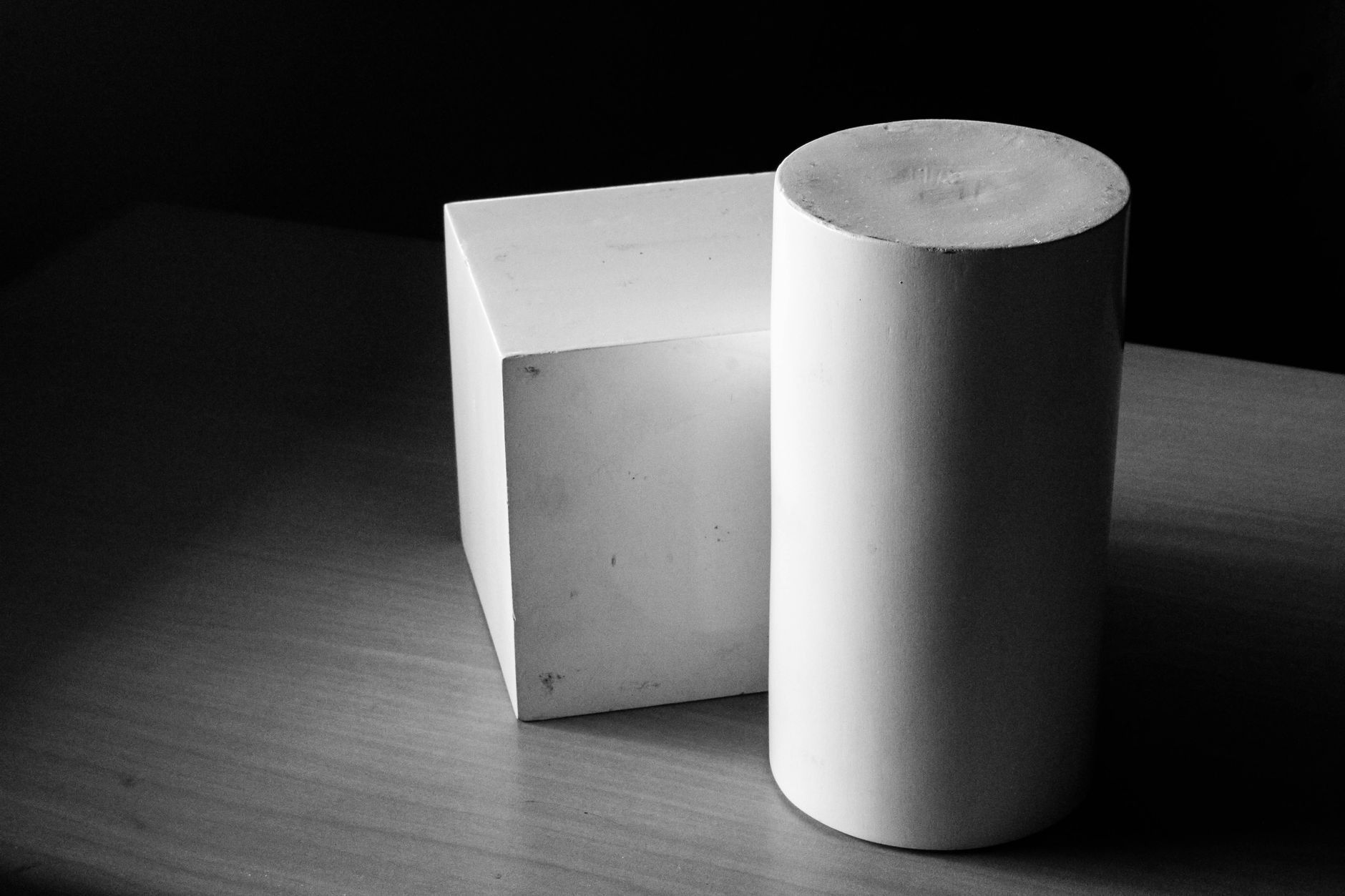 Black and white photo of a cube and cylinder on a wooden table, showcasing geometric forms.